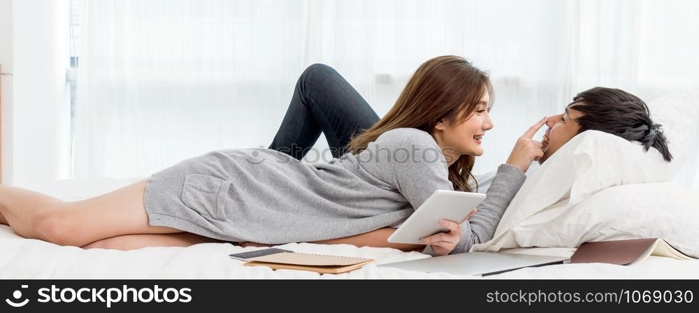 Banner and cover scene of Happy Asian couple talking and smiling with technology tablet and laptop on the bedroom at home, young women pointing to the nose of boyfriend, Lover and life style concept,