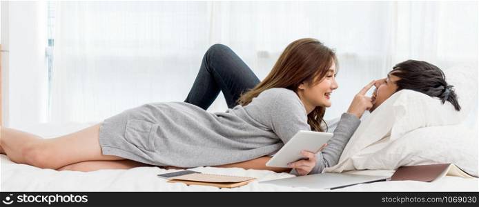 Banner and cover scene of Happy Asian couple talking and smiling with technology tablet and laptop on the bedroom at home, young women pointing to the nose of boyfriend, Lover and life style concept,