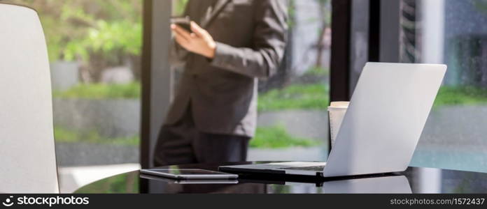 Banner of Closeup technology laptop and tablet with paper coffee cup on the working space in modern office over the photo blurred of asian businessman standing and using mobile phone background
