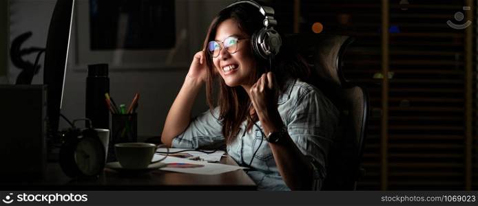 Banner, web page or cover template of Portrait of Asian Businesswoman listening the music via headphone and smart mobile phone with on table in workplace at late time, Work hard and too late concept