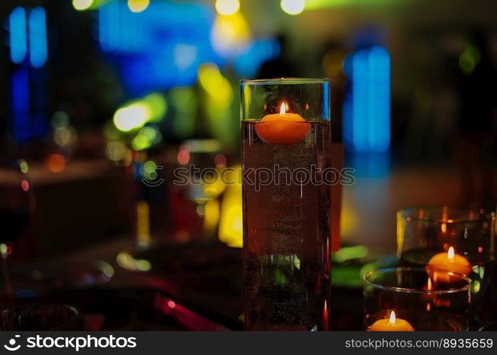 Banquet table decorated with burning candles in glass vases in restaurant hall. In the background party with silhouettes of people dancing on the dance floor with disco lights glowing searchlight.. Banquet table decorated with burning candles in glass vases in restaurant hall. In the background party with silhouettes of people dancing on the dance floor with disco lights glowing searchlight