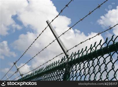 Barbed wire on top of chain security fence against blue cloudy sky.