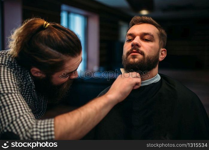 Barber combing beard of the client man in salon cape at the barbershop