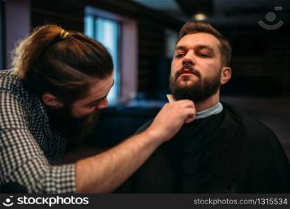 Barber combing beard of the client man in salon cape at the barbershop