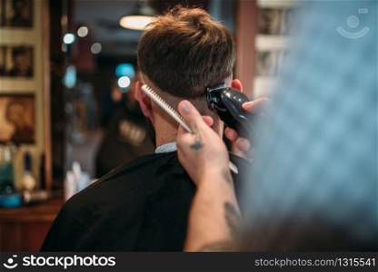 Barber hands makes hairstyle of the client man by clipper. Male person sitting at the mirror in barbershop