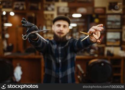 Barber in hat and gloves shows sciccors. Professional barbershop is a trendy occupation. Male hairdresser in retro style hairdressing salon. Barber shows sciccors, retro style barbershop