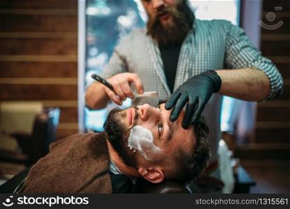 Barber shaves the beard of the client by straight razor at barbershop.. Barber shaves beard of the client at barbershop