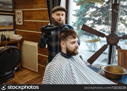 Barber with comb and hairdryer makes a hairstyle to a client. Professional barbershop is a trendy occupation. Male hairdresser and customer in retro style hair salon. Barber with comb and hairdryer makes a hairstyle