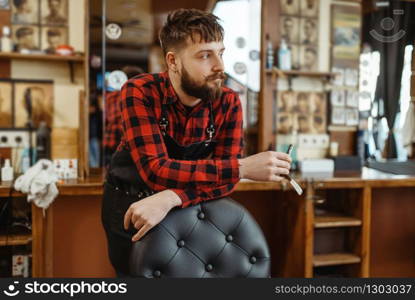 Barber with razor blade poses at the chair. Professional barbershop is a trendy occupation. Male hairdresser in retro style hair salon, accessories for cutting. Barber with razor poses at the chair, barbershop
