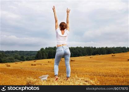 barefoot girl with straw hat stands on a haystack on a bale in the agricultural field after harvesting.. girl with straw hat stands on a haystack on a bale in the agricultural field after harvesting