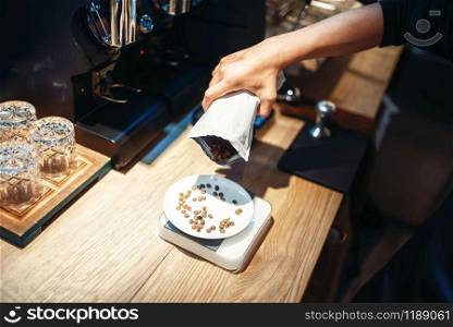 Barista hand pours coffee beans into the plate standing on scales, wooden counter on background. Professional espresso preparation by bartender in cafeteria. Barista hand pours coffee beans into the plate