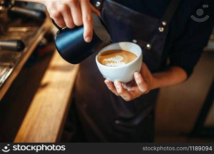 Barista hand pours cream into the cup of coffee, wooden table on background. Professional cappuccino preparation by bartender. Barista hand pours cream into the cup of coffee