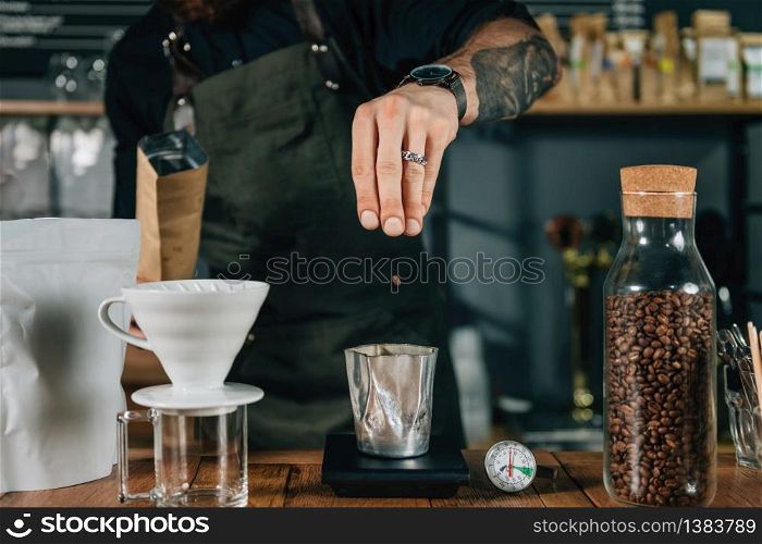 Barista hands pouring fresh roasted coffee beans for weighing on digital scale. Tools and equipment for making Drip Brew coffee on wooden table. Barista with tattooed arms wearing dark uniform.