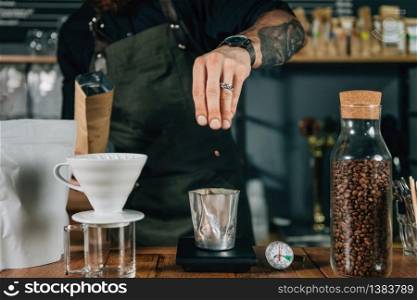 Barista hands pouring fresh roasted coffee beans for weighing on digital scale. Tools and equipment for making Drip Brew coffee on wooden table. Barista with tattooed arms wearing dark uniform.