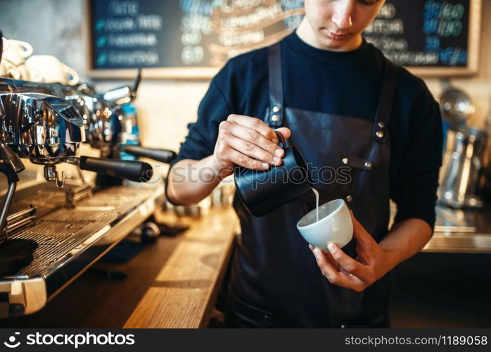 Barista in apron pours cream into the cup of coffee, cafe counter on background. Professional cappuccino preparation by bartender. Barista pours cream into the cup of coffee