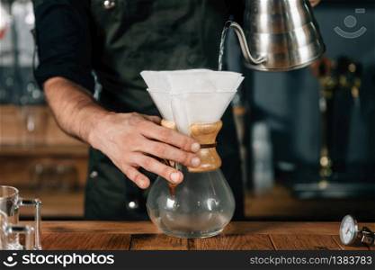 Barista pouring boiling water from kettle into white filter for filter coffee. Barista with tattooed arms wearing dark uniform.