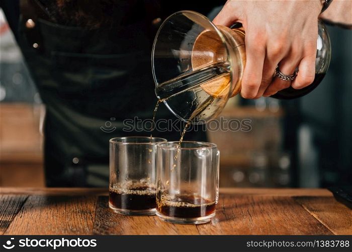 Barista pouring two Chemex coffees. Tools and equipment for making Chemex coffee on table. Barista with tattooed arms wearing dark uniform.