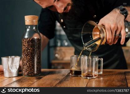 Barista pouring two filter coffees. Tools and equipment for making filter coffee on table. Barista with tattooed arms wearing dark uniform.