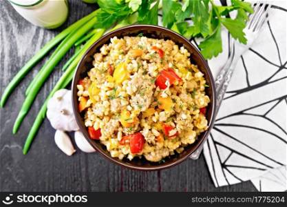 Barley porridge with minced meat, yellow and red bell peppers, garlic and onions in a clay bowl, a towel and parsley on wooden board background from above