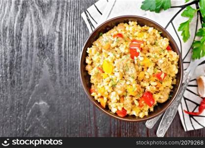 Barley porridge with minced meat, yellow and red bell peppers, garlic and onions in a clay bowl, napkin and parsley on wooden board background from above