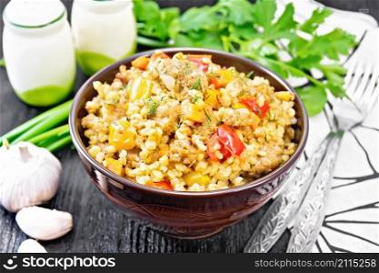 Barley porridge with minced meat, yellow and red bell peppers, garlic and onions in a clay bowl, a towel and parsley on black wooden board background