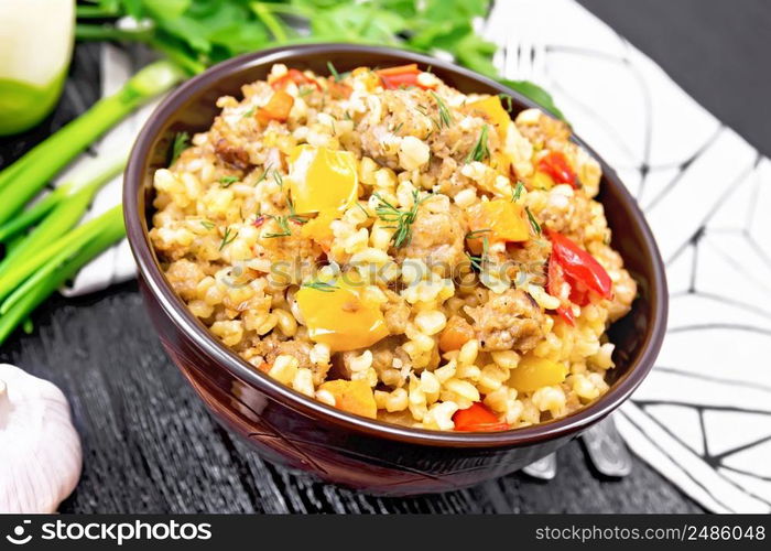 Barley porridge with minced meat, yellow and red bell peppers, garlic and onions in a clay bowl, napkin and parsley on wooden board background