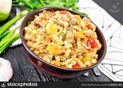 Barley porridge with minced meat, yellow and red bell peppers, garlic and onions in a clay bowl, napkin and parsley on wooden board background