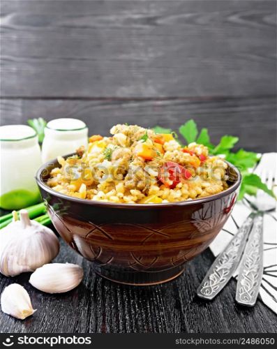Barley porridge with minced meat, yellow and red bell peppers, garlic and onions in a clay bowl, a towel and parsley on wooden board background