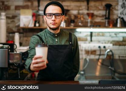 Barman in black apron stretches plastic cup of coffee in hands.. Barman in black apron stretches plastic cup