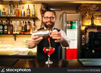 Barman is making alcohol cocktail at bar counter. Barman with shaker and glass of beverage. Barman is making alcohol cocktail at counter