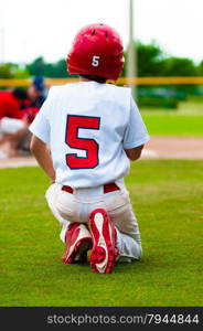 Baseball boy kneeling on the ground while an injured player is on the field.