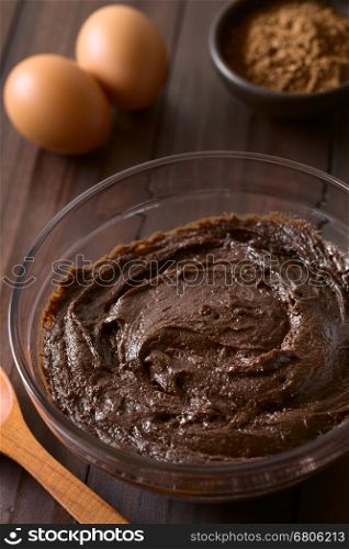 Basic homemade brownie, chocolate cake or cookie dough in glass bowl with cocoa powder and eggs in the back, photographed on dark wood with natural light (Selective Focus, Focus one third into the image)