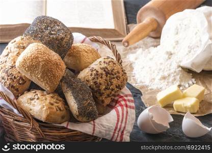 Basket with healthy homemade bread rolls decorated with seeds, surrounded by flour, butter, eggs and an open cookbook, on a black wooden table.