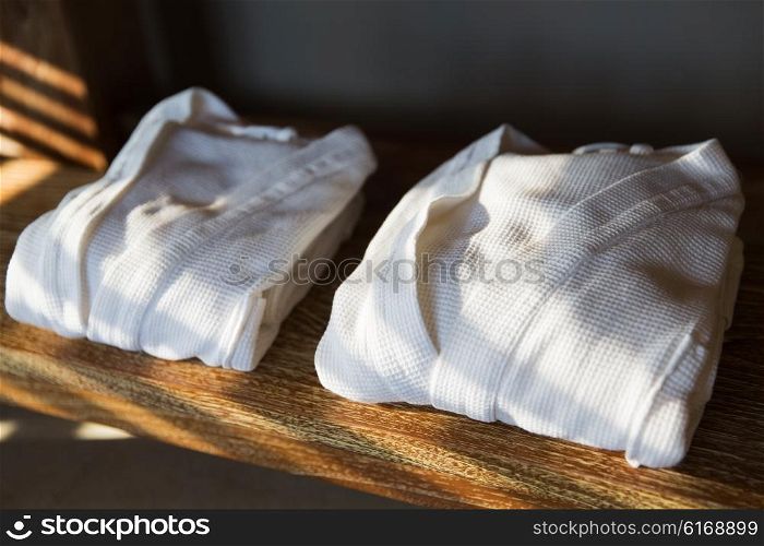bath, clothes, hygiene and luxury concept - close up of two white bathrobes on wooden shelf at home or hotel
