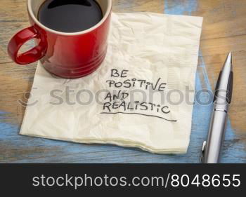 be positive and realistic - handwriting on a napkin with a cup of coffee