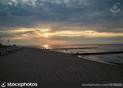Beach at Kuehlungsborn in the evening with people strolling and the beach chairs on the left edge and the typical groynes that protrude into the water. The sun shines through the clouds in the sky. Beach at Kuehlungsborn in the evening