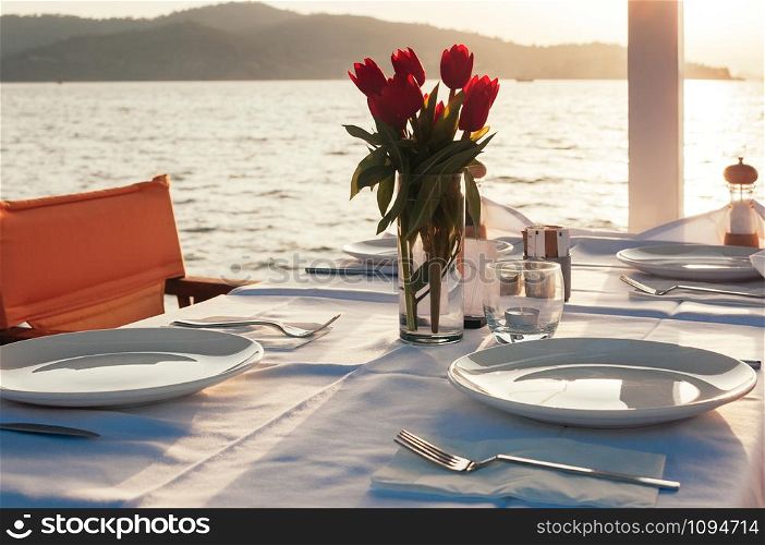 Beach restaurant with sea view at sunset, a table setting decorated with flowers. Empty porcelain plates on cloth. Beautiful summer holiday concept. Romantic dinner at resort. Selective focus