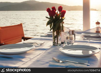 Beach restaurant with sea view at sunset, a table setting decorated with flowers. Empty porcelain plates on cloth. Beautiful summer holiday concept. Romantic dinner at resort. Selective focus