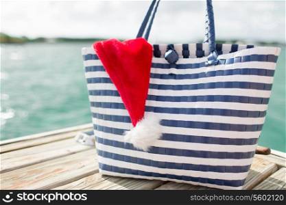 beach, summer, vacation, christmas and accessories concept - close up of beach bag and santa helper hat on wooden pier