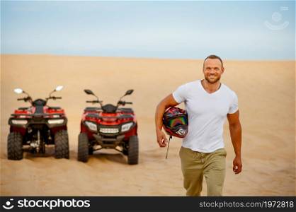 Bearded atv racer holds helmet in hand, desert sands on background, quadbike racer. Male person on quad bike, sandy race on quadbike, dune safari in hot sunny day, 4x4 extreme adventure, quad-biking. Bearded atv racer, desert sands on background