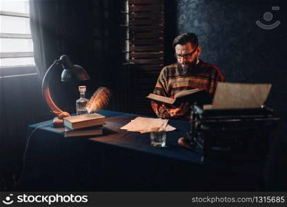 Bearded author in glasses reading a book. Retro typewriter, feather, crystal decanter, books and vintage lamp on the desk. Bearded author in glasses reading a book