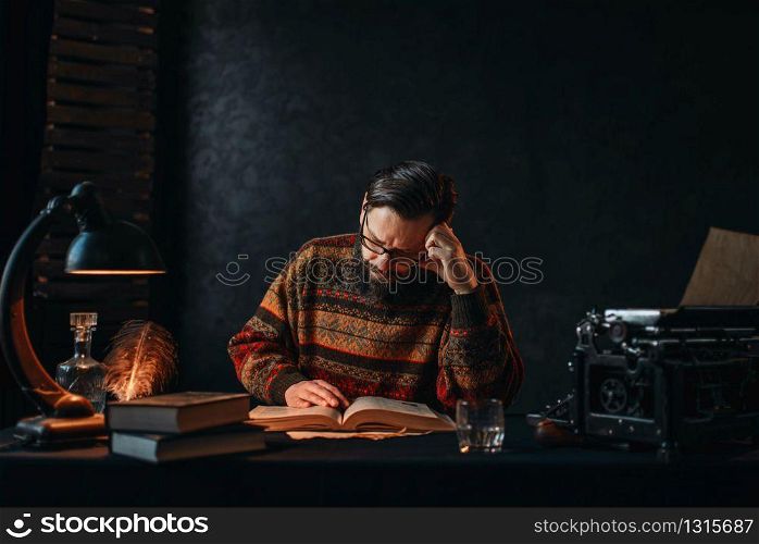Bearded author in glasses reading a book. Retro typewriter, feather, crystal decanter, books and vintage lamp on the desk. Bearded author in glasses reading a book