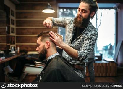 Bearded barber cutting male client hairstyle by scissors. Man in black salon cape at the barbershop