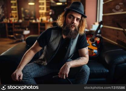 Bearded barber sitting on black leather couch, guitar and barbershop inerior on background