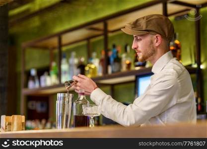 Bearded barman in white shirt and cap pouring liquor into shaker while preparing cocktail on counter in pub. Male barkeeper pouring alcohol into shaker