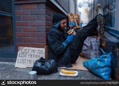 Bearded beggar eats burger on city street. Poverty is a social problem, homelessness and loneliness, alcoholism and drunk addiction, urban lonely. Bearded beggar eats burger on city street