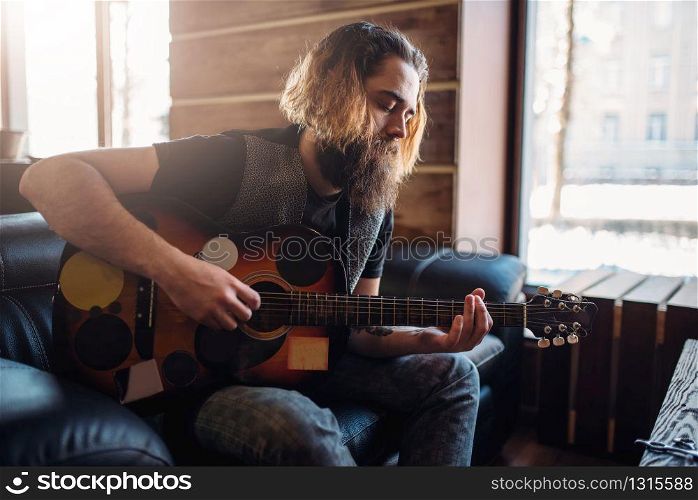 Bearded brutal man with guitar sitting on black couch. Barber shop inerior on background