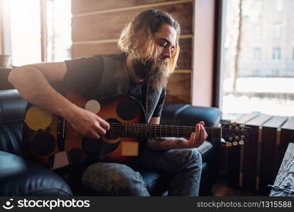 Bearded brutal man with guitar sitting on black couch. Barber shop inerior on background