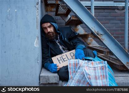 Bearded bum with help sign on city street. Poverty is a social problem, homelessness and loneliness, alcoholism and drunk addiction, urban lonely. Bearded bum with help sign on city street