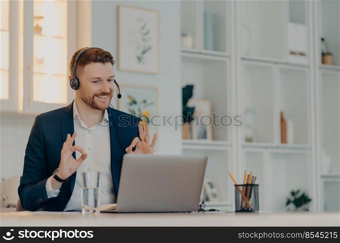 Bearded businessman very happy with result of online negotiations while using laptop and headset, happy male office worker showing ok sign with both hands and smiling. Business and work concept. Happy executive manager showing ok sign while talking online